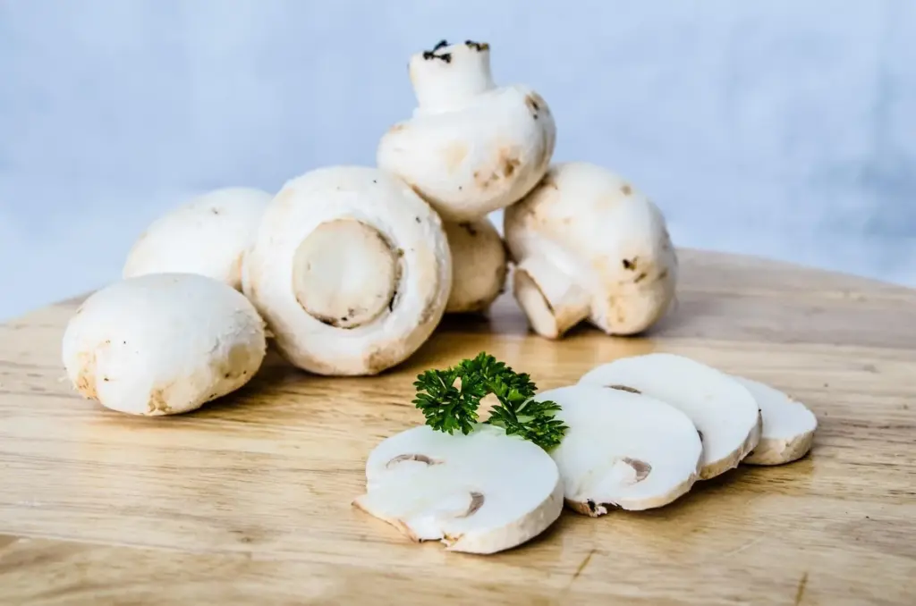 Funghi champignon freschi, interi e affettati, su un tagliere in legno con un rametto di prezzemolo