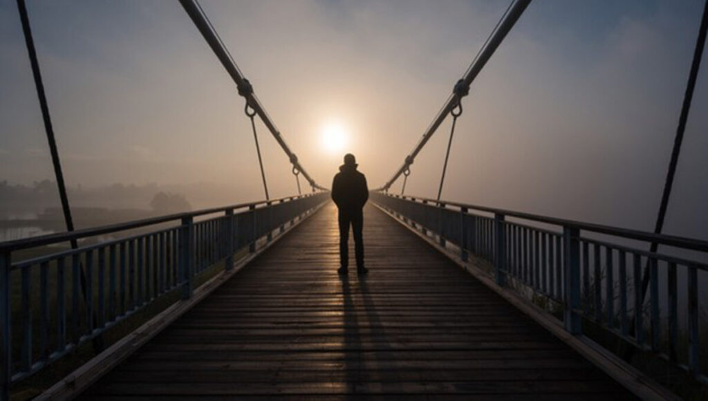 Viaggiatore in silhouette su un ponte sospeso avvolto dalla nebbia al sorgere del sole