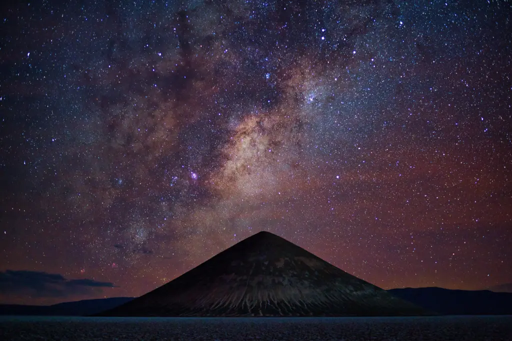 Il nucleo della Via Lattea con sfumature dorate e rossastre sopra la sagoma di una montagna a forma di cono, in un cielo notturno stellato