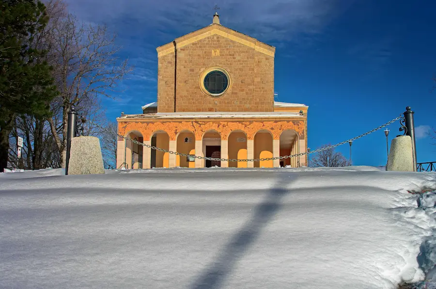 Il Santuario di Monte delle Formiche innevato, con la facciata in pietra e il porticato affrescato sotto un cielo blu intenso