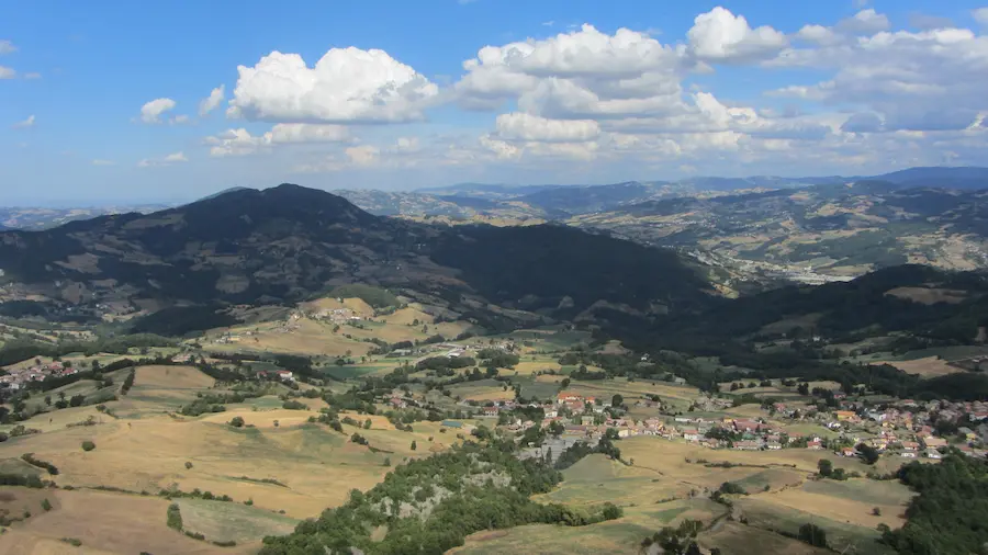 Veduta aerea su un borgo appenninico circondato da campi e boschi, con dorsali montuose sullo sfondo sotto un cielo con nuvole cumuliformi
