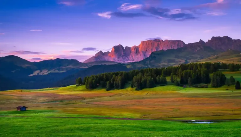 Paesaggio montano al tramonto con prati verdi e montagne illuminate dalla luce rosa.