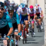Gruppo di ciclisti professionisti in gara lungo una strada, con un corridore in primo piano e il plotone alle sue spalle.