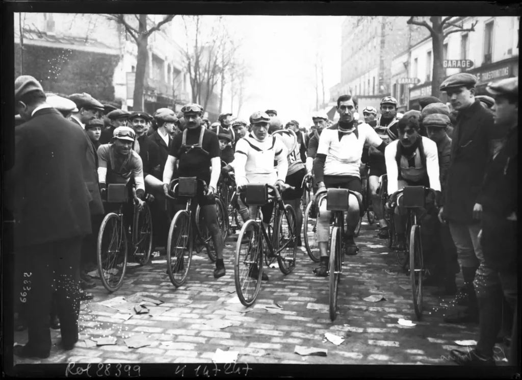 Partenza della Paris–Tours 1913 a Suresnes, con i ciclisti della squadra Française-Hutchinson sulle biciclette d’epoca, circondati dal pubblico